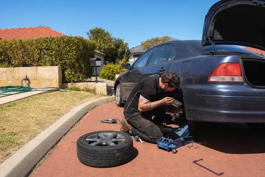 Man kneeling at a rear wheel with the tyre off, inspecting the brake assembly in a driveway