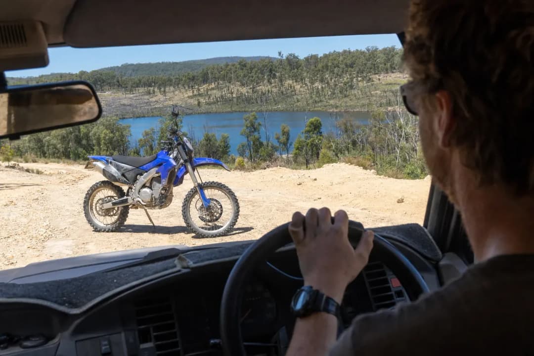 Blue dirt bike parked on sandy terrain with a forest and lake in the background.