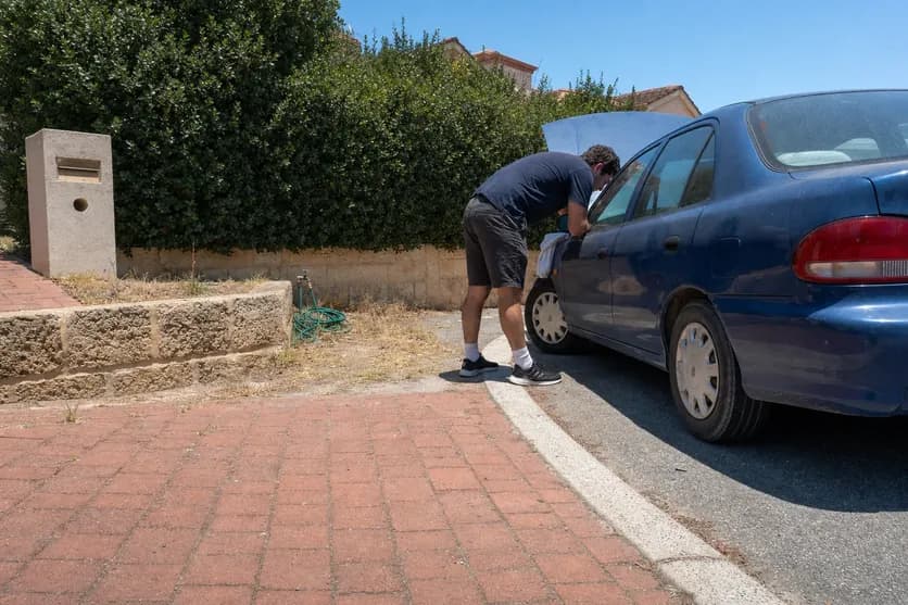 Driver leaning under the open bonnet of a blue sedan parked at the kerb