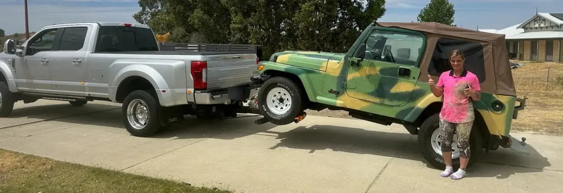 Customer gives a thumbs up beside her camouflage Jeep Wrangler on the Quokka Towing wheel lift, back home on her driveway