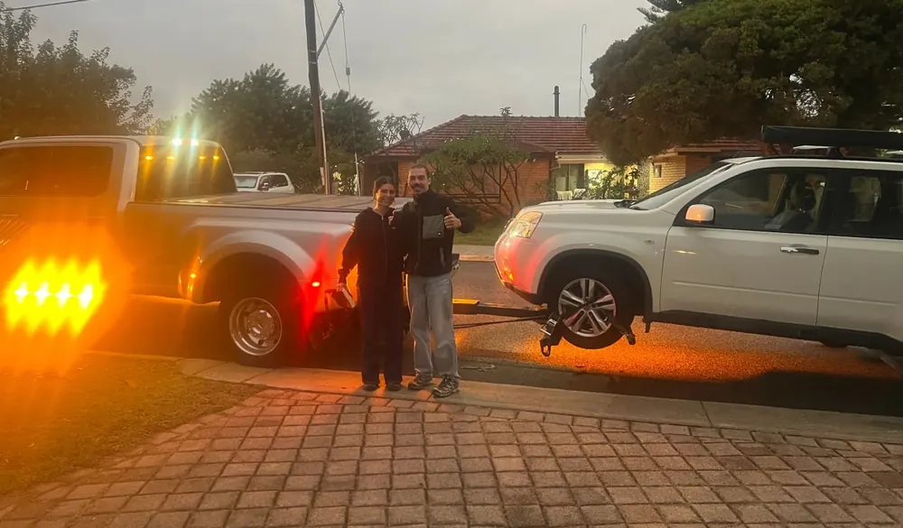 The two customers give a thumbs up beside their white Nissan X-Trail on the Quokka Towing wheel lift at dusk