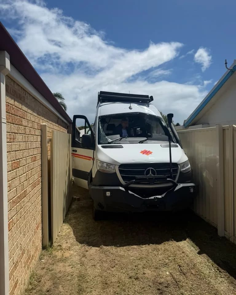 The white Sprinter van in the narrow side passage between the house and the fence, with the driver's door open