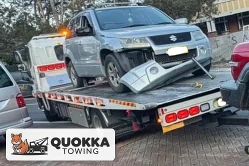 Silver Suzuki Grand Vitara with front-end crash damage strapped onto a Quokka Towing tilt tray on a Perth street