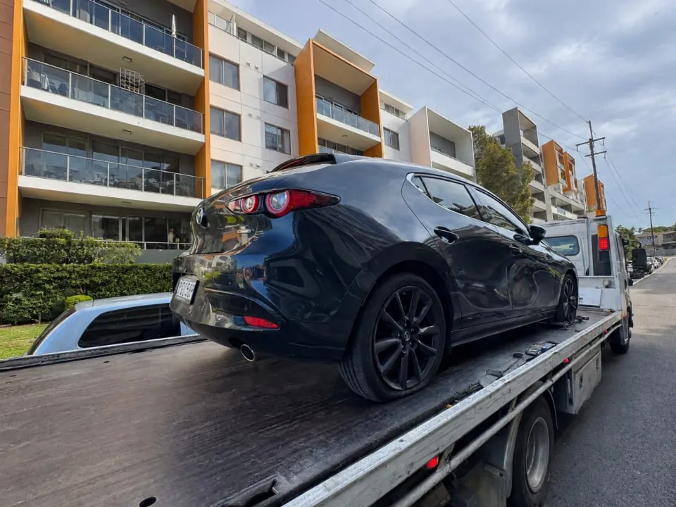Quokka Towing tilt tray carrying a black hatchback past an apartment block