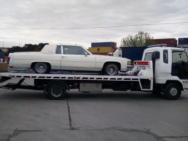 Tilt tray carrying a white classic car past a container yard