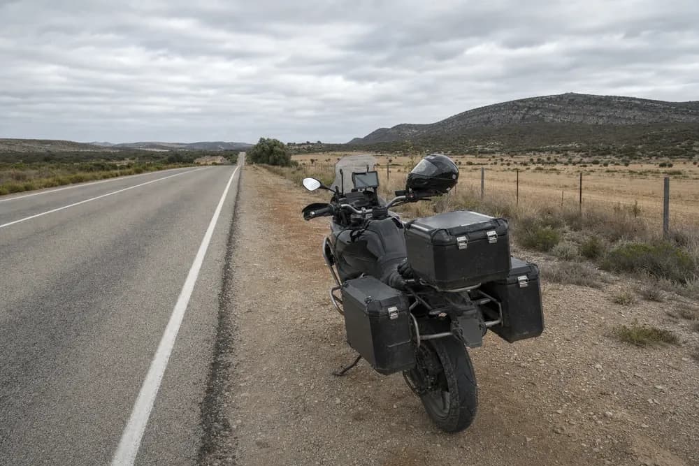 Black touring motorcycle with panniers and helmet parked beside a rural road under a cloudy sky.